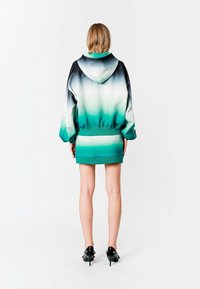 Sudadera con capucha en degradado de negro a azul verdoso, con un ajuste relajado, dobladillo acanalado y detalles gráficos. Combinada con una falda corta en azul verdoso.
