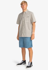 Giovane uomo in piedi che indossa una camicia a maniche corte beige a quadri, pantaloni corti in velluto blu e scarpe alte nere con lacci bianchi.