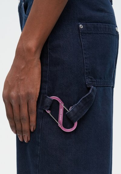 Denim overal s tmavomodrou tkaninou, ktorý má výrazný ružový kovový karabín pripevnený k slučke, čím zdôrazňuje funkčnosť a dizajnové detaily.