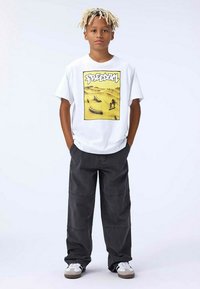 Hvid grafisk t-shirt med et gult rektangel, der har teksten "FREEDOM" samt en ørken scene. Sammen med løstsiddende mørkegrå bukser.