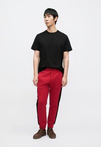 Punaiset collegehousut mustilla sivuraitoisilla, yhdistettynä mustaan T-paitaan. Malli käyttää ruskeita lenkkareita. Yksivärinen tausta neutraaleissa väreissä.