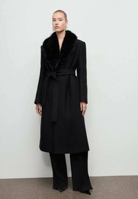 Abrigo negro largo con un cuello de pelo sintético texturizado, cinturón en la cintura y silueta recta, combinado con pantalones negros de pierna ancha y zapatos con tacón.
