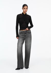 Haut noir à manches longues avec un design enveloppant et une texture transparente, associé à un jean gris taille haute et large. Des talons noirs complètent la tenue.