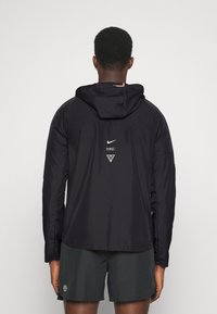 Czarna kurtka Nike z kapturem, z logo i trójkątami na plecach, wykonana z gładkiego, lekkiego materiału.