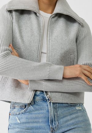 Grijze cropped jack met geribbelde grijze mouwen, een opgestikte kraag, een ritssluiting aan de voorkant en zijzakken, gecombineerd met blauwe distressed jeans.
