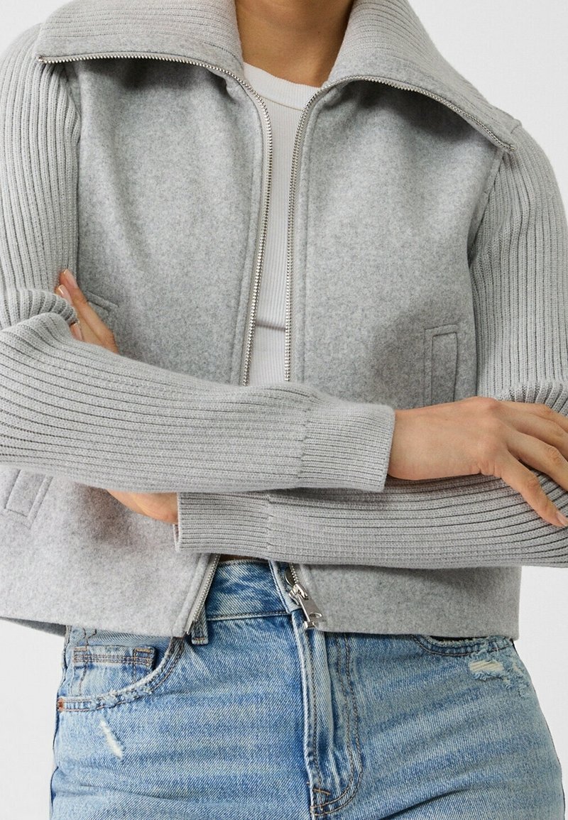 Gray cropped jacket with ribbed gray sleeves, a folded collar, front zip, and side pockets, paired with blue distressed jeans.