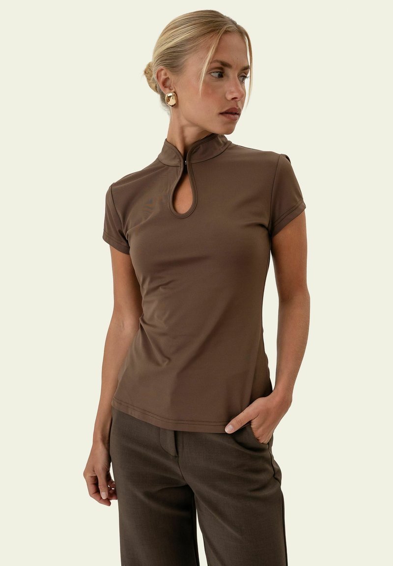 Femme aux cheveux blonds portant un haut marron à manches courtes avec une ouverture en forme de trou de serrure à l'avant et un pantalon marron, posant avec une main dans la poche.
