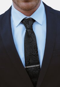 Corbata negra con estampado de paisley y textura suave, llevada con una camisa de color azul claro, debajo de una chaqueta de traje oscura. Presenta un clip de corbata plateado como detalle.