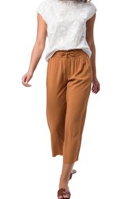 Mujer vestida con una blusa blanca de encaje floral, pantalones cortos marrones claros con cordón y sandalias marrones caminando hacia adelante sobre un fondo blanco.