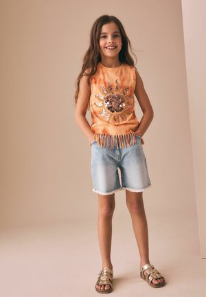 Girl smiles wearing sleeveless orange fringe top with sequined sun and light denim shorts standing against beige background.