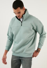 Sudadera ligera de color teal con cuello alto, cierre de media cremallera en la parte delantera, puños acanalados y dobladillo. Presenta una textura suave, similar al algodón.