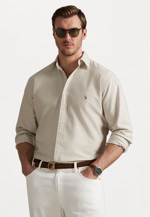 Beige knappad skjorta med långa ärmar, bärs med vita byxor och ett brunt bälte. Gröna solglasögon och en klocka fulländar outfiten.