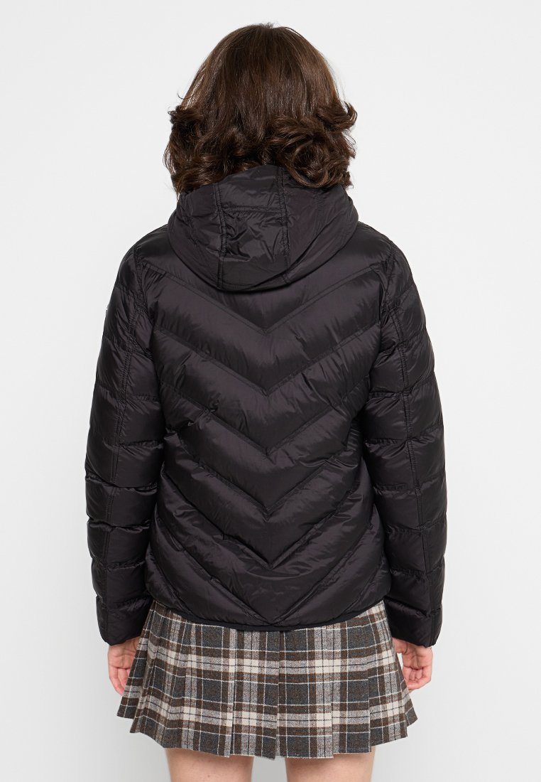 Doudoune noire avec un motif en losanges, capuche fixe et silhouette ajustée. Portée sur une jupe à carreaux.