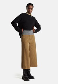 Schwarzer Strickpullover über einem grauen gerippten Strickbund, kombiniert mit weiten beigen Cordhosen und schwarzen Schnürkampfschuhen.