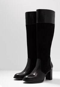 Bottes noires montant jusqu'au genou avec un bout et un talon en cuir lisse, dotées d'une tige en daim doux et d'un bord en cuir lisse. Talon bloc.