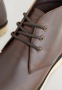 Brown leather shoe with dark brown laces threaded through three brass eyelets, detailed stitching, and a cream-colored sole visible.