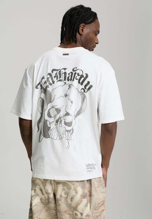 Hombre con el cabello trenzado que lleva una camiseta blanca de Ed Hardy con un gran diseño en la espalda de una calavera agrietada y una serpiente.