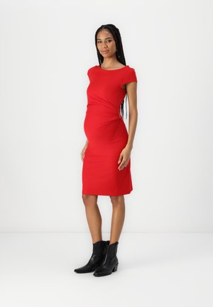 Rotes Umstandskleid mit figurbetontem Schnitt, kurzen Ärmeln und gerafftem Taillendetail. Getragen mit schwarzen Stiefeletten. Glatte Textur.