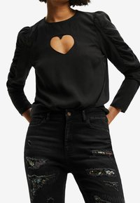 Blouse noire à manches longues avec une découpe en forme de cœur au niveau de la poitrine. Éléments de manches froncées et texture lisse. Associée à un jean noir orné d'une broderie colorée et d'accents en détresse.
