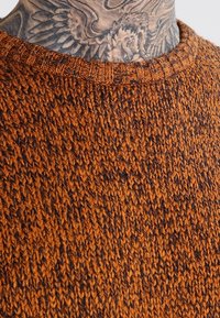 Pull en tricot de couleur orange chaud avec un motif texturé et un col côtelé, montrant des variations subtiles dans la teinte du fil.