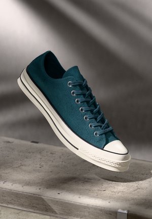 Teal suede sneaker with a rubber sole, white toe cap, and dark laces. Features textured upper and contrasting white stripe along the sole.
