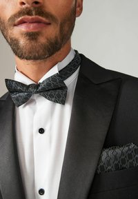 Black bow tie with a textured pattern, paired with a white shirt and a black suit jacket, featuring a matching pocket square with a similar design.