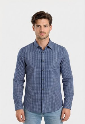 Hombre con camisa azul ajustada de manga larga con patrón y botones, y jeans azul claro, de pie contra un fondo blanco liso.