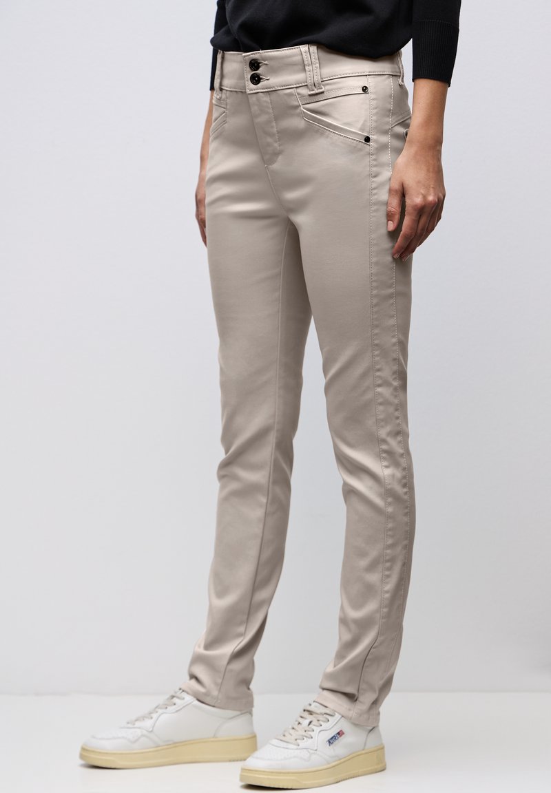 Beżowe spodnie o dopasowanym kroju z zapięciem na guzik i zamek, noszone z białymi tenisówkami i czarną bluzką z długim rękawem, stojące na białej podłodze.