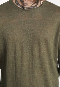 Olivegrünes Langarmshirt aus leichtem Stoff, mit rundem Halsausschnitt und leicht durchscheinender Textur. Minimalistisches Design ohne Muster.