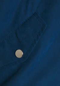 Niebieska tkanina z przeszywaną klapą z zabezpieczeniem na pojedynczy okrągły srebrny guzik zatrzaskowy, ukazująca szczegółową fakturę i linie szwów.