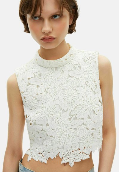 Jeune personne portant un haut court en dentelle florale blanche sans manches avec un col montant et un ourlet en festons, associé à un jean en denim clair.