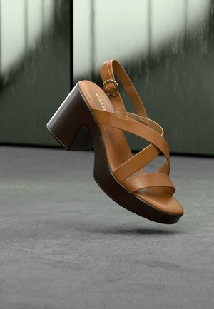 Sandale compensée en cuir marron avec talon bloc et bride à la cheville, suspendue en l'air au-dessus d'un sol gris sur un fond sombre flou.