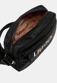 Borsa a tracolla nera in nylon con chiusura a zip, dotata di un logo rosa chiaro sulla parte anteriore e un interno beige a motivo. Strap regolabile incluso.
