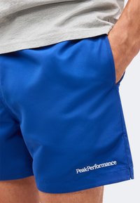 Άνδρας που φορά φωτεινά μπλε σορτς με το λογότυπο "PeakPerformance" στην τσέπη του αριστερού ποδιού, χέρι μέσα στην τσέπη και ανοιχτό γκρι πουκάμισο.