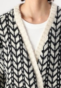 Cardigan a motivi houndstooth in bianco e nero con colletto in maglia color crema, caratterizzato da una consistenza morbida e un design aperto davanti.