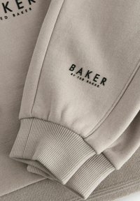 Sweatshirt beige avec des poignets côtelés, présentant un détail textuel noir "BAKER" sur le côté. Texture douce et design minimaliste.