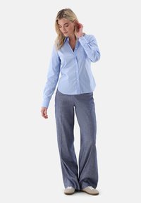 Light blue, button-up shirt with a pocket and long sleeves, paired with gray, wide-leg trousers and white sneakers, against a plain background.