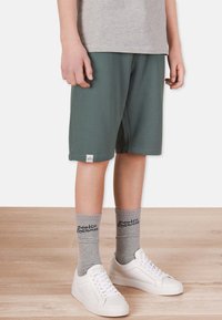 Gröna shorts i mjukt material med elastisk midja. Grå strumpor med text och vita sneakers. Bakgrund med trämönstrad golv.