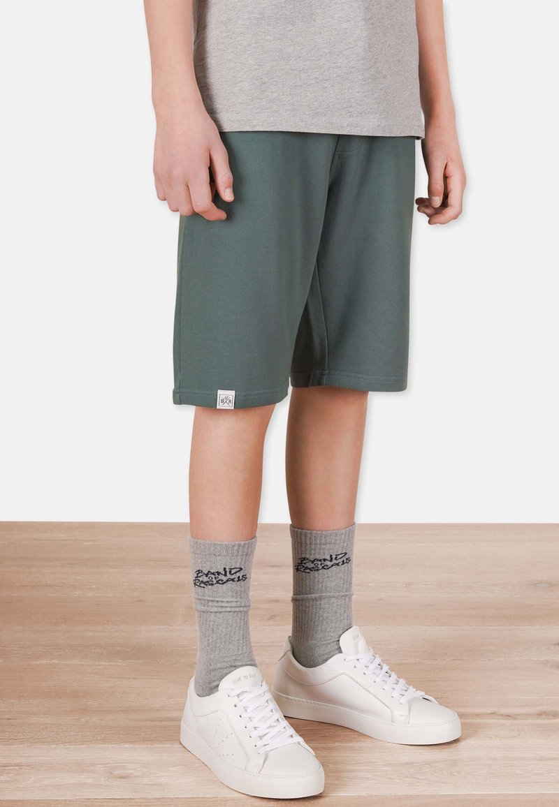 Gröna shorts i mjukt material med elastisk midja. Grå strumpor med text och vita sneakers. Bakgrund med trämönstrad golv.