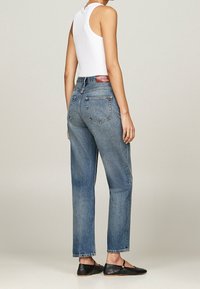 Vaqueros de mezclilla azul de tiro alto con pernera recta, que cuentan con una etiqueta de cuero en la parte trasera y detalles sutiles de costura. Combinados con una camiseta sin mangas blanca.