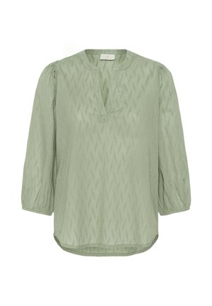 Svetlo zelena bluzica z teksturo, s krilnimi rokavi dolžine tri četrtine, okroglim ovratnikom z V-rezom in rahlo ukrivljenim robom.