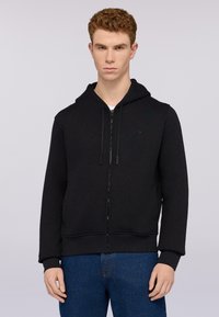 Černá mikina na zip vyrobená z hladké tkaniny, s kapucí na šňůrku a předními kapsami, doplněná modrými džíny.