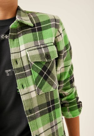 Homme portant une chemise en flanelle à carreaux verts, noirs et blancs par-dessus un t-shirt noir à motif, avec la manche de la chemise remontée au-dessus du coude.