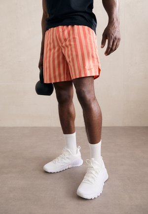 Osoba w pomarańczowych, prążkowanych szortach i białych sneakersach trzyma w jednej ręce czarny kettlebell na jednolitym tle.