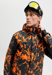 Chaqueta de esquí con un patrón negro y naranja, cuello alto, capucha ajustable y bolsillos. Textura suave y diseño moderno.
