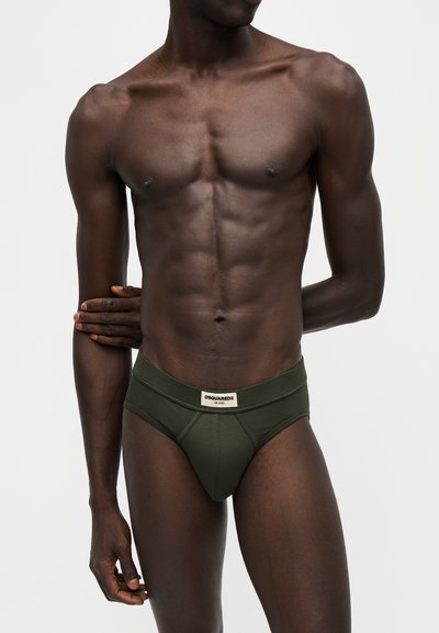 Torse masculin musclé à la peau foncée portant un slip vert Dsquared2, les mains croisées, sur un fond gris clair uni.