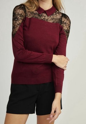 Kobieta ubrana w bordowy sweter z czarną koronką na ramionach i kołnierzykiem, w połączeniu z czarnymi szortami, z założonymi na wysokości talii skrzyżowanymi ramionami.