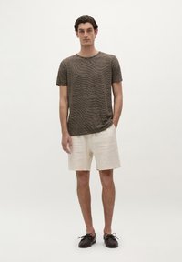 Camiseta de manga corta a rayas marrones, pantalones cortos de lino beige y zapatos oscuros. El modelo está de pie contra un fondo claro, con las manos en los bolsillos.
