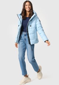 Lichtblauwe gewatteerde jas met capuchon, ritssluiting en zwarte accenten. Gecombineerd met blauwe high-waisted jeans en beige sneakers.