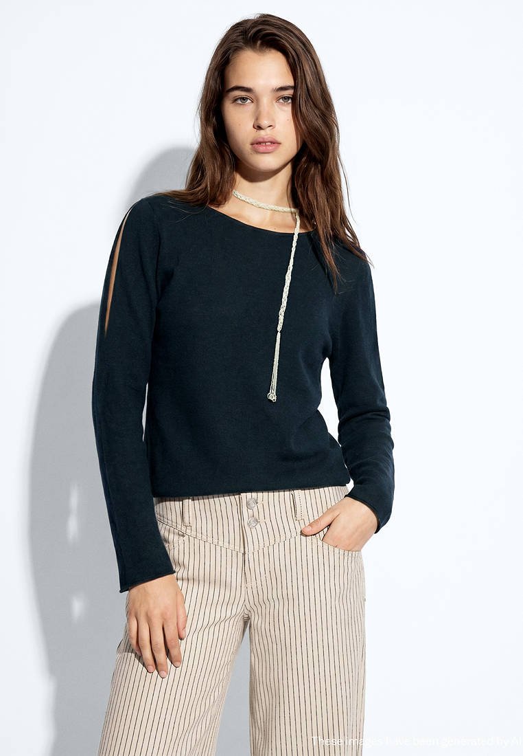 Jeune femme aux longs cheveux bruns portant un haut noir à manches longues avec des fentes aux manches et un pantalon beige rayé taille haute, debout devant un fond blanc.